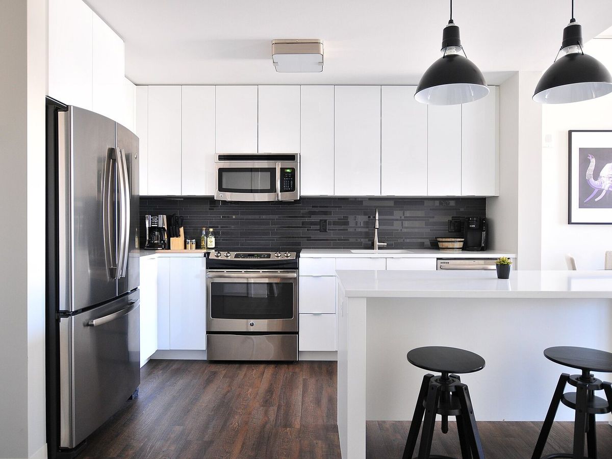 A white kitchen with stainless steel appliances, dark wood floors, and an island with two stools under black pendant lights