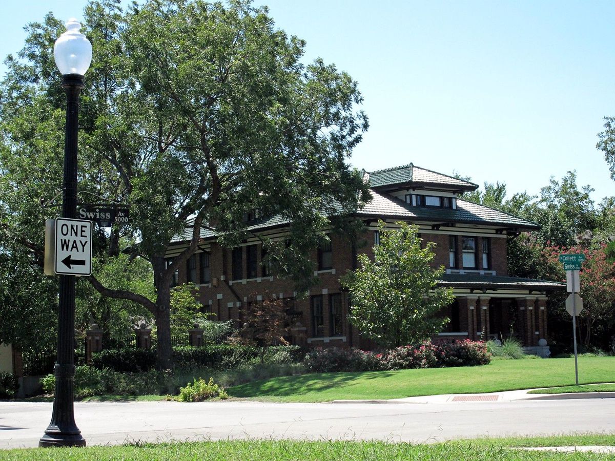 An early 1900s brick Prairie-style house with wide eaves behind a corner lawn on Swiss Avenue