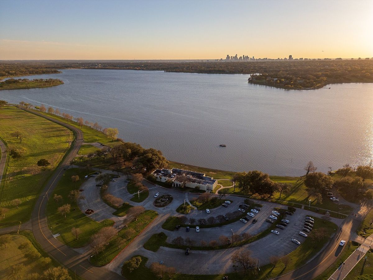 Aerial view of White Rock Lake at sunset with the Bath House Cultural Center on the shore and the Dallas skyline in the distance